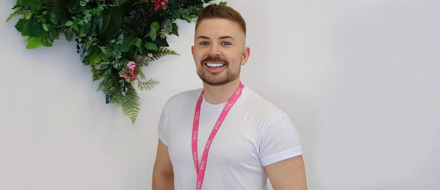 Luke Manton, owner of Manton Executives, stands smiling against a white wall with a decorative arrangement of green foliage and pink flowers. He is wearing a white t-shirt and a pink lanyard with "ME" branding