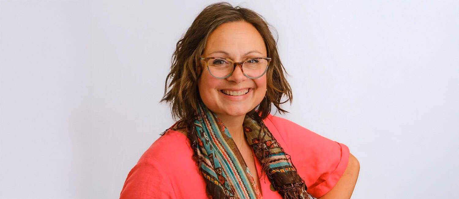Dr Nancy Doyle, Founder of Genius Within CIC, sits smiling in a bright office space. She has wavy hair and is wearing glasses and a brightly coloured dress
