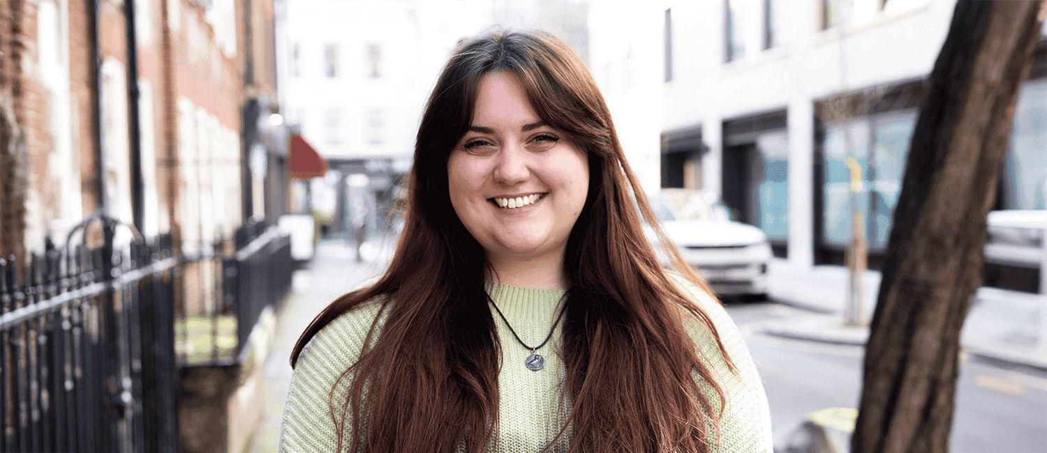 A young woman stands outdoors, smiling and wearing a light green jumper and a necklace, with urban buildings and a tree in the background.