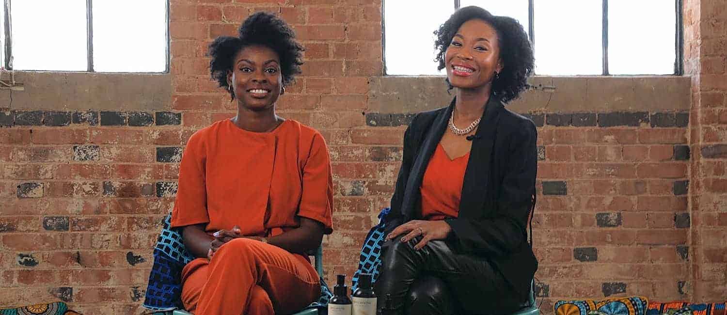 Rachael Twumasi-Corson and Joycelyn Mate, co-founders of Afrocenchix, a Black-owned natural haircare brand, sit smiling in a creative space with product bottles displayed in front of them