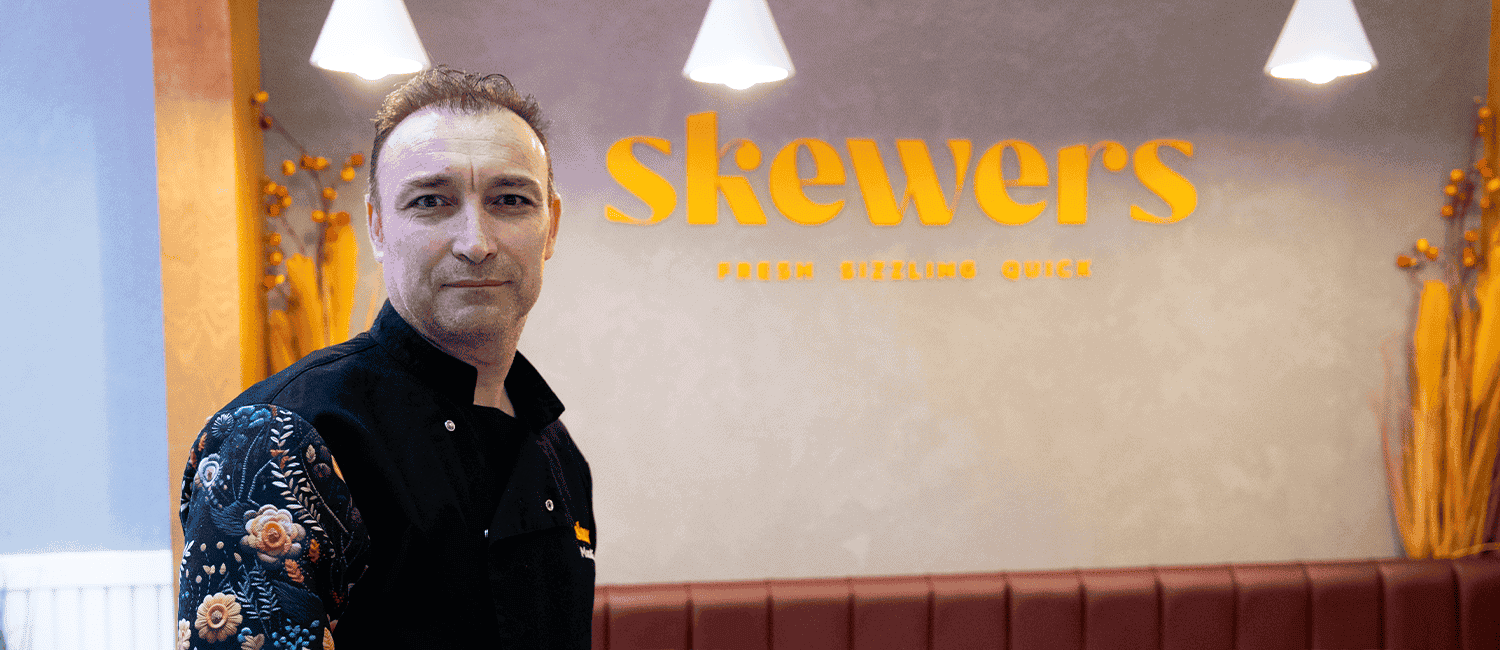 A close-up shot of Head Chef of Skewers, Murat, in his chef's uniform, standing to the left in front of the restaurant's branded wall that displays 'Skewers' in orange lettering