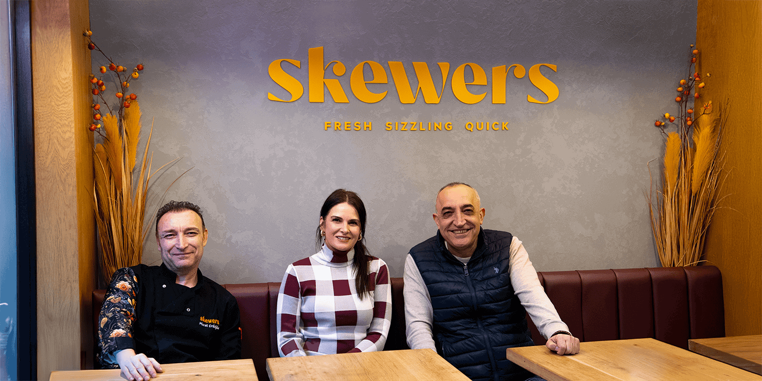 Business owners and Head Chef of Skewers, sitting at a table inside their restaurant, against a grey wall with the orange branded lettering 'Skewers' on the wall, above the slogan 'Fresh, Sizzling, Quick'