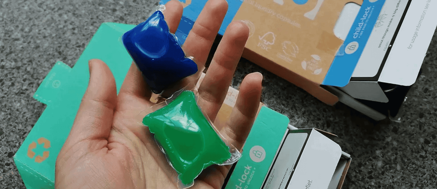 An open hand holding one blue and one green laundry capsule in its palm. One blue and one green small cardboard box can be seen below the hand.