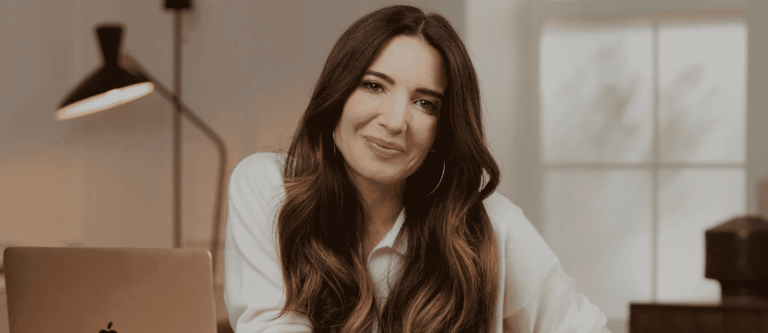 A woman with long brown hair and hoop earrings smiles warmly, sitting in a cosy room with a laptop. Soft lighting and a lamp create a calm atmosphere.