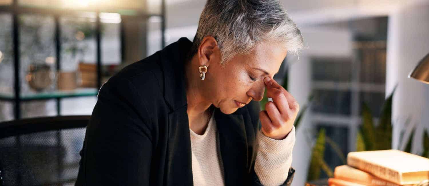 Businesswoman sitting at a desk, appearing tired and stressed, with eyes closed and pinching the bridge of her nose in a modern office setting