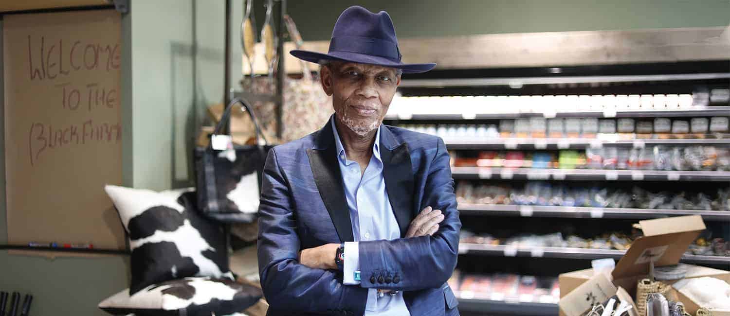 Wilfred Emmanuel-Jones MBE, founder of The Black Farmer, a premium Black-owned food brand, stands with arms crossed in a grocery store, wearing a stylish blue suit and hat