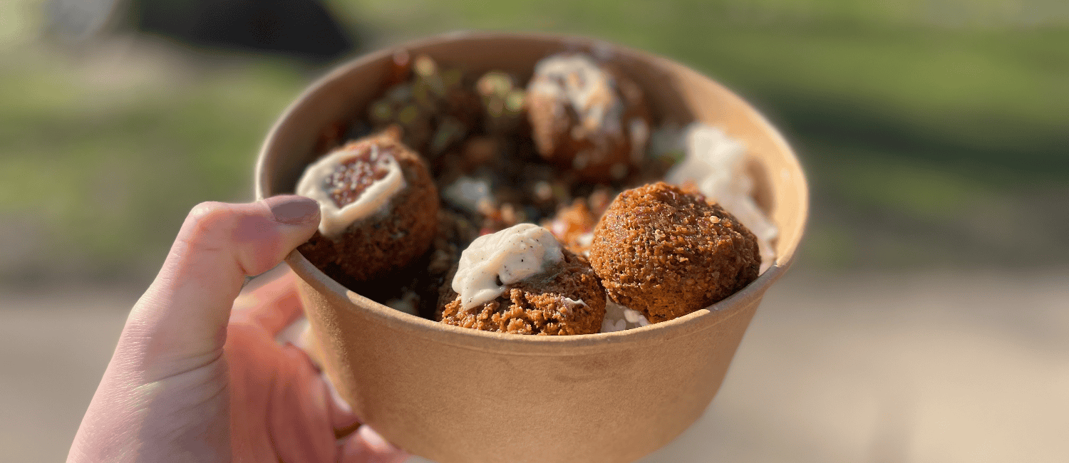 Close-up shot of a brown cardboard bowl of falafel and healthy grains, with a light dressing of sauce and a green, blurred backdrop
