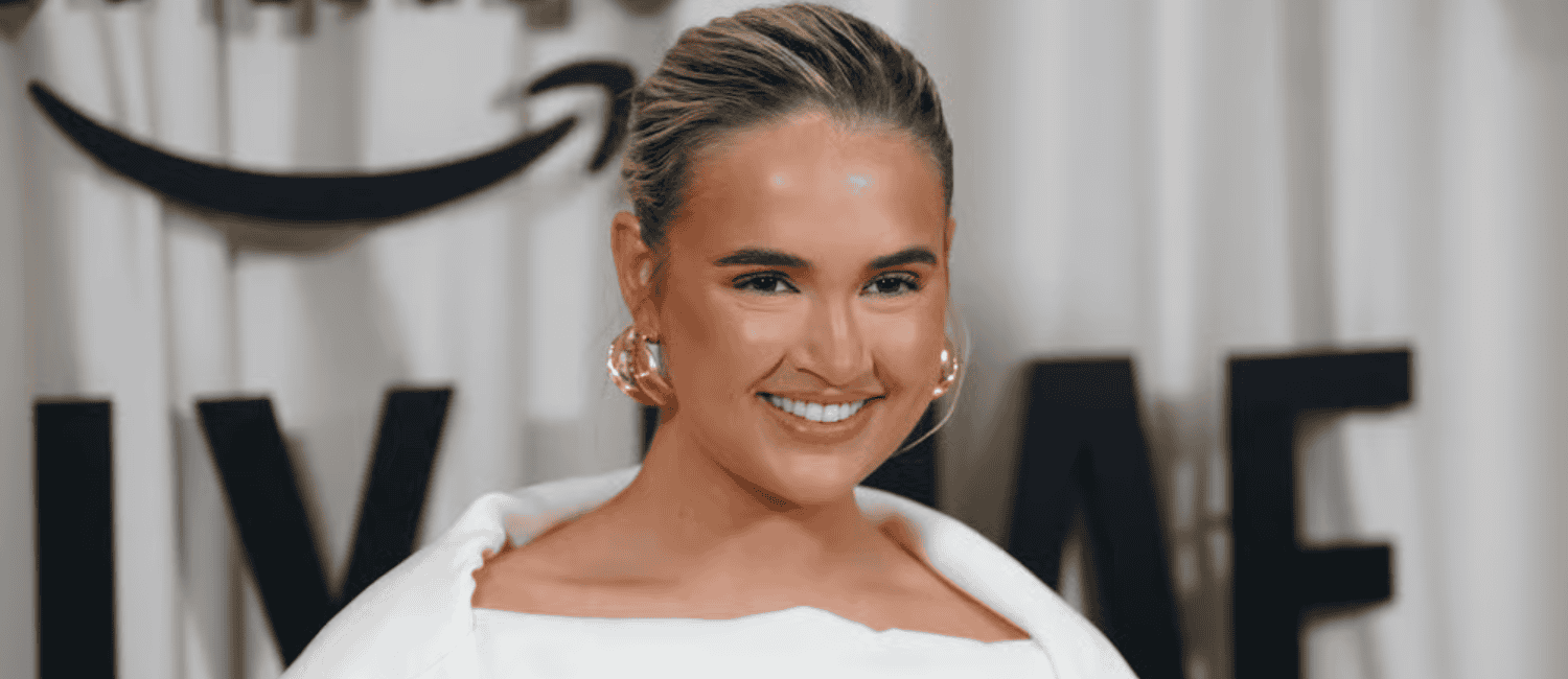 Molly-Mae Hague smiles at the camera, wearing large gold hoop earrings and a white off-the-shoulder outfit. A blurred backdrop with black text and an Amazon logo is visible behind her