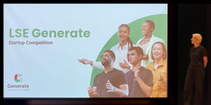Presenter standing beside large screen displaying LSE Event about startup competition with diverse speakers.
