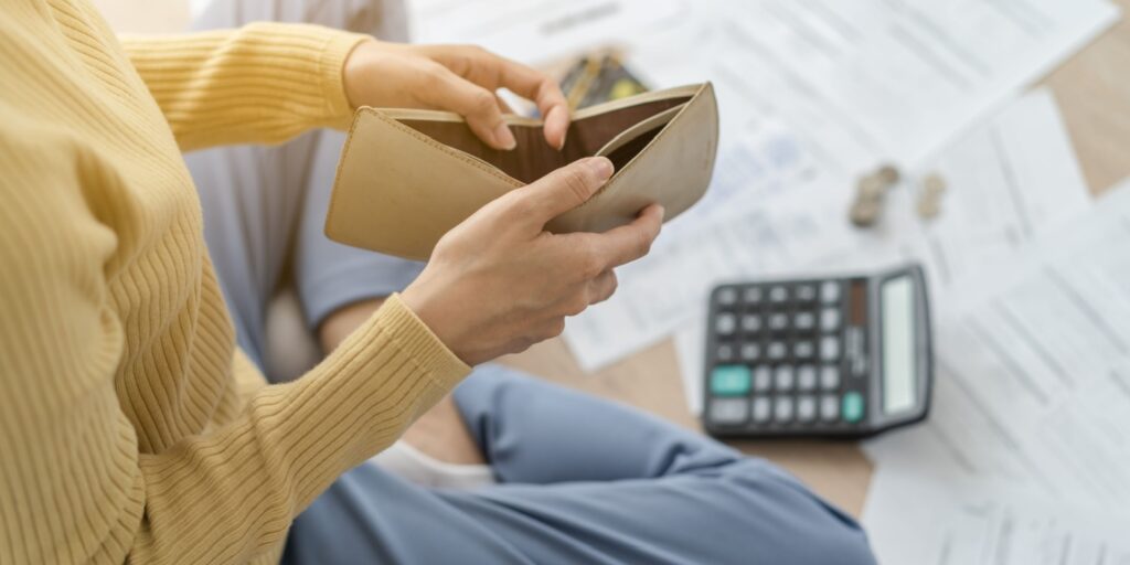 Person holding an empty wallet with financial documents and a calculator on the floor, showing the challenge of trying to start a business with no money