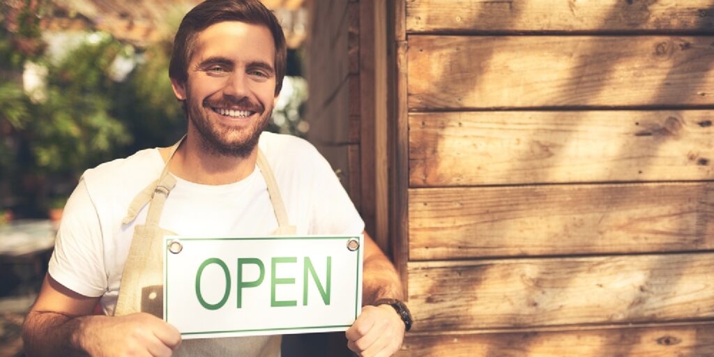Portrait of a smiling coffee shop owner holding an OPEN sign outside his business.