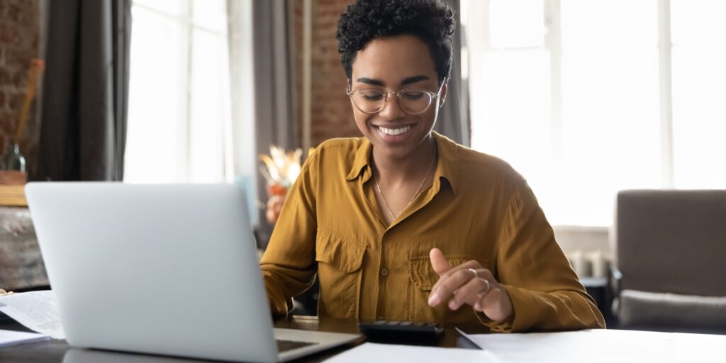 Happy young female business owner in glasses, working with a calculator at a laptop computer, with large windows in the background.