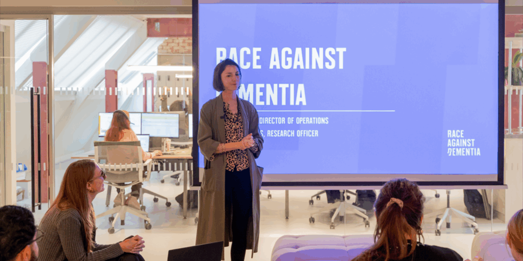 Lydia Beaton giving a presentation to a seated audience in an office setting, with a large screen behind her displaying the title "Race Against Dementia." The talk is focused on the organisation’s work, presented to staff at 1st Formations