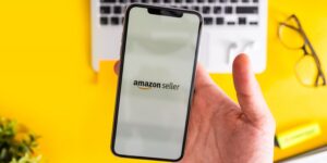 Person holding a smartphone displaying the Amazon Seller app on screen, with a laptop and glasses in the background, highlighting the decision Amazon FBA sellers face when choosing to operate as a sole trader or limited company.