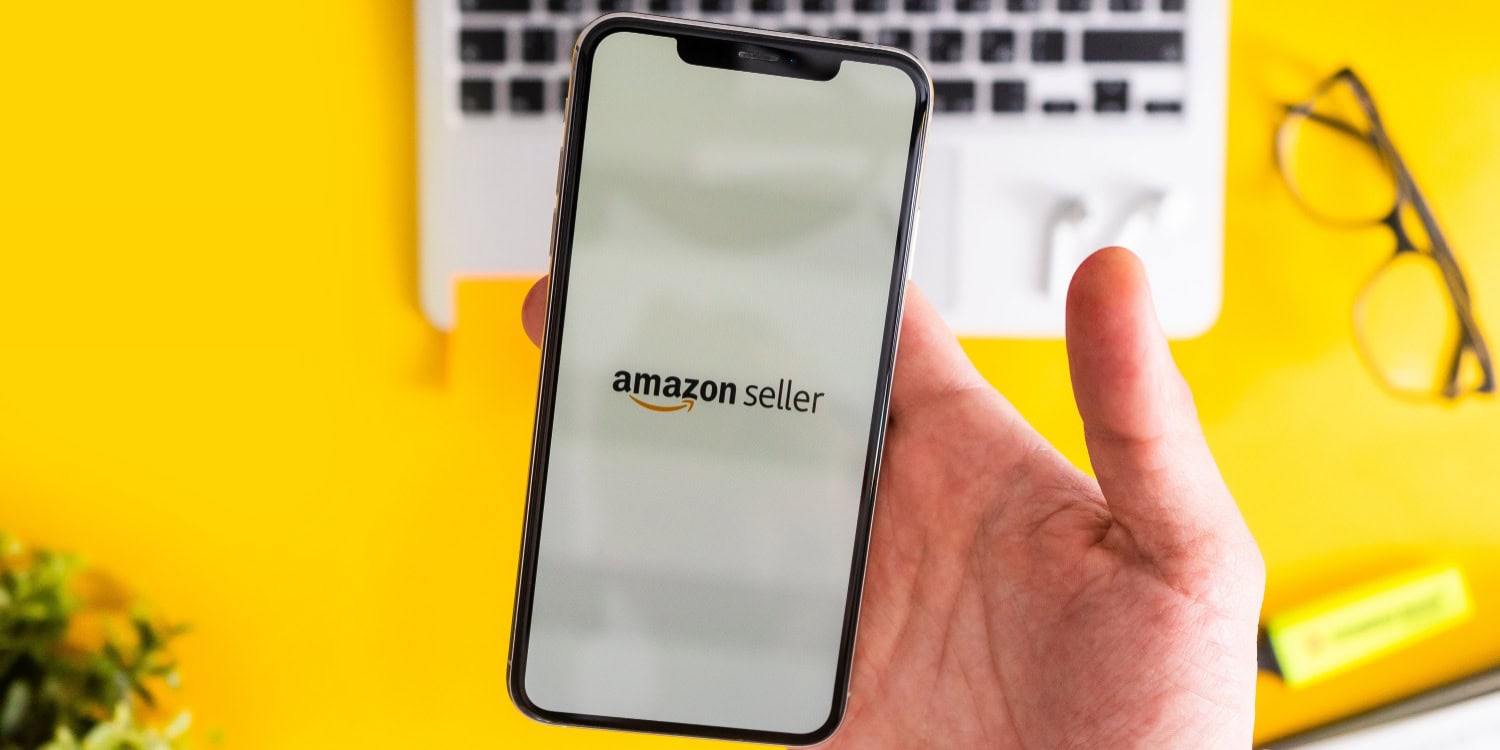 Person holding a smartphone displaying the Amazon Seller app on screen, with a laptop and glasses in the background, highlighting the decision Amazon FBA sellers face when choosing to operate as a sole trader or limited company.