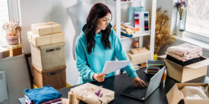 Entrepreneur working on laptop surrounded by fabrics, packages, and products - turning side hustle into small business