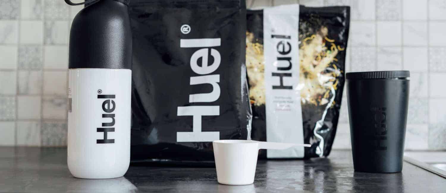 Huel branded nutrition products including a white bottle, black tumbler, measuring scoop, and black and white food pouches on a kitchen counter