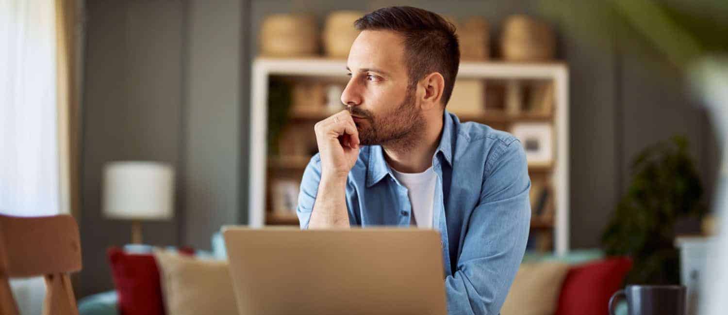 Entrepreneur sitting at a desk with a laptop, resting his chin on his hand and looking thoughtfully into the distance in a cosy home office setting