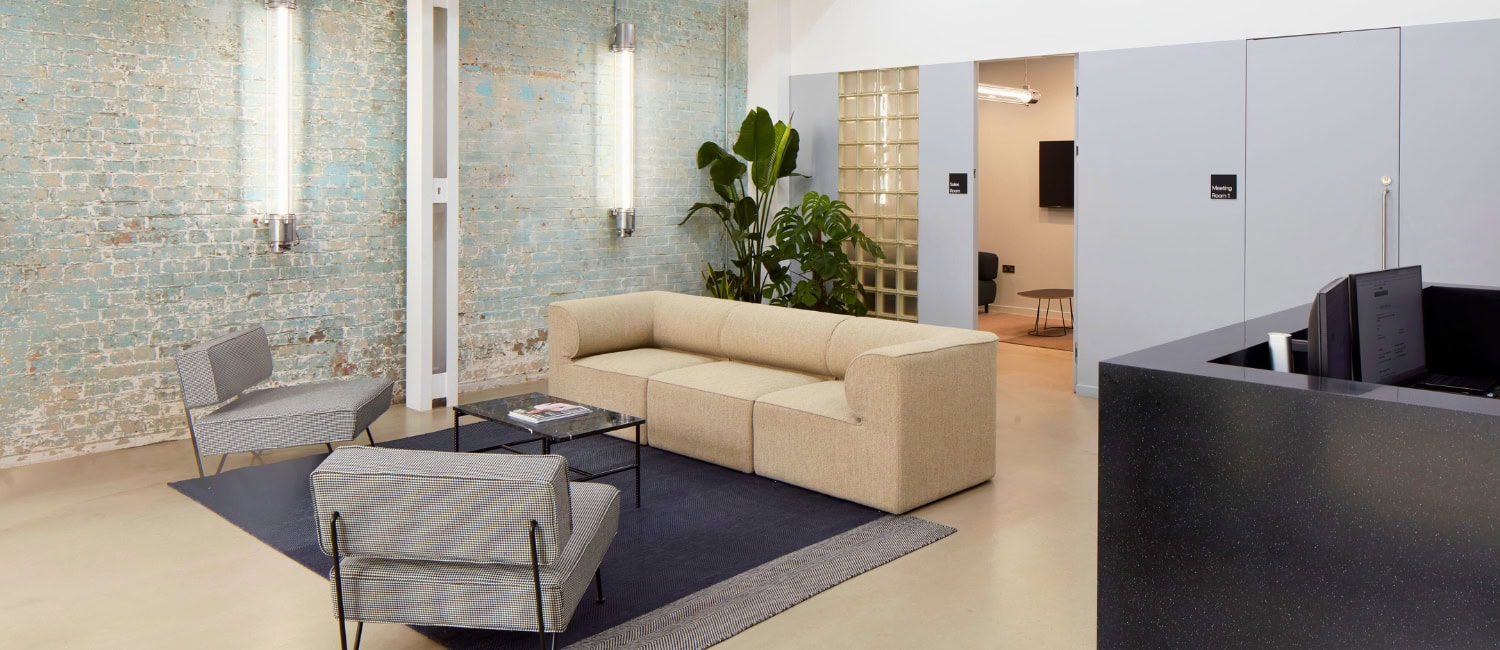 Modern office lobby with industrial decor, featuring a beige sofa, gray chairs, exposed ductwork, and a brick accent wall.