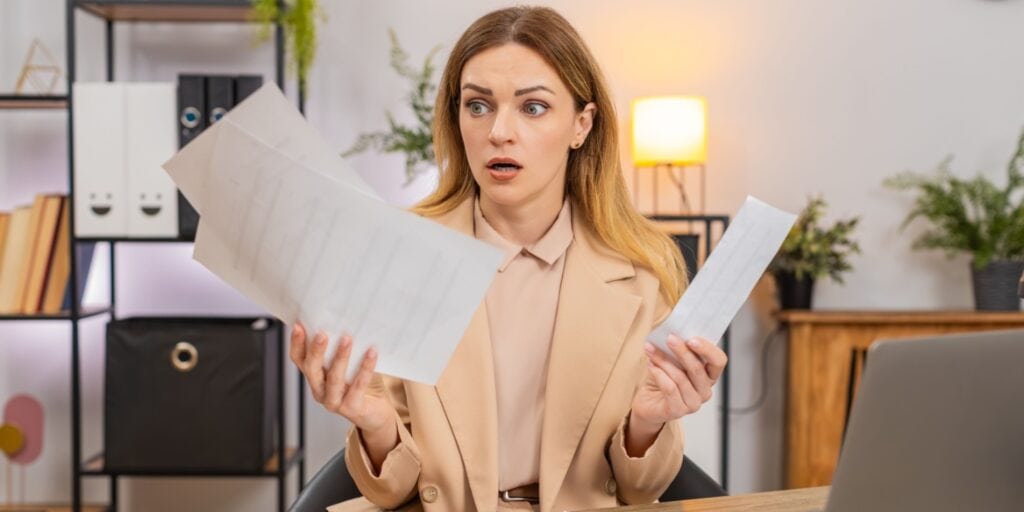 A surprised woman discovering hidden costs when running her business, dressed in business attire and sitting at a desk holding several sheets of paper, looking shocked as she reviews unexpected documents.
