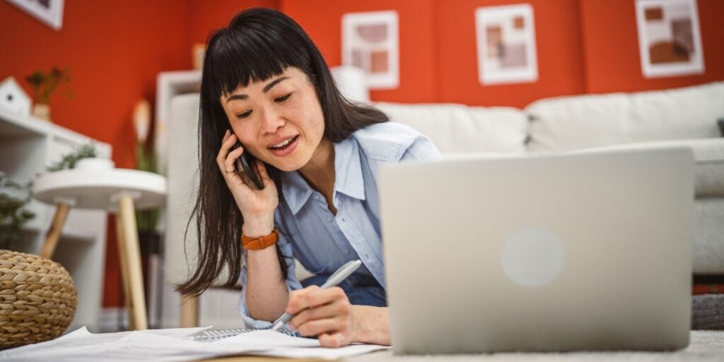 Woman multitasking at home, talking on the phone while writing notes and working on a laptop.