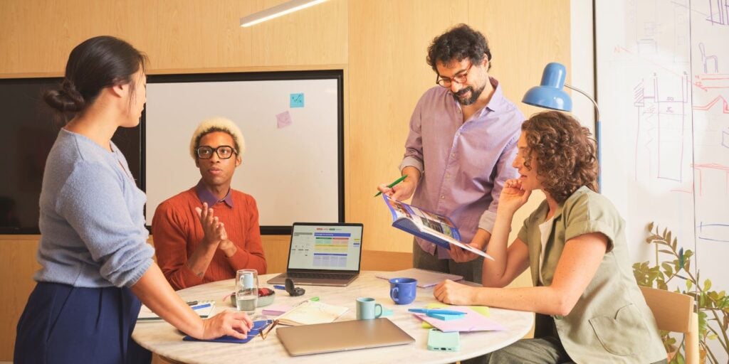 Four colleagues collaborating in a modern office meeting, reviewing documents and discussing ideas around a table with a laptop and notes.