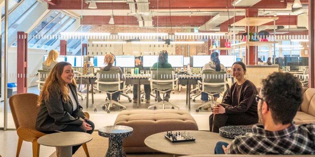 Three colleagues converse in a lounge area with a chessboard on the table; behind them a glass wall reveals a busy open‑plan office with rows of monitors.