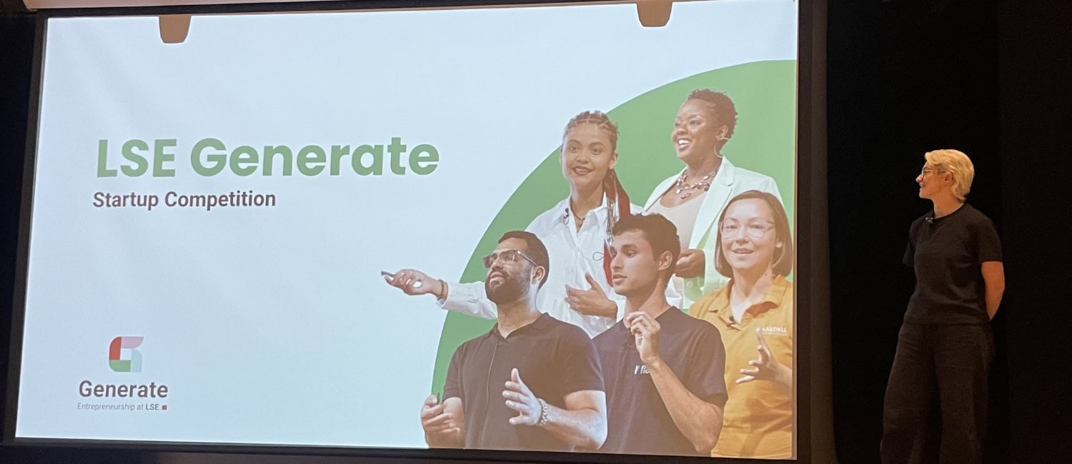 Presentation slide featuring the title "LSE Generate Startup Competition" alongside images of diverse individuals engaged in discussion. A presenter stands to the right, looking at the screen.