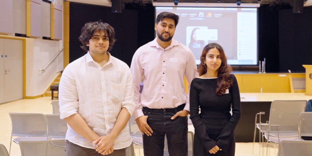Three students pose for the camera during a presentation, with a digital display in the background showing a virtual participant.