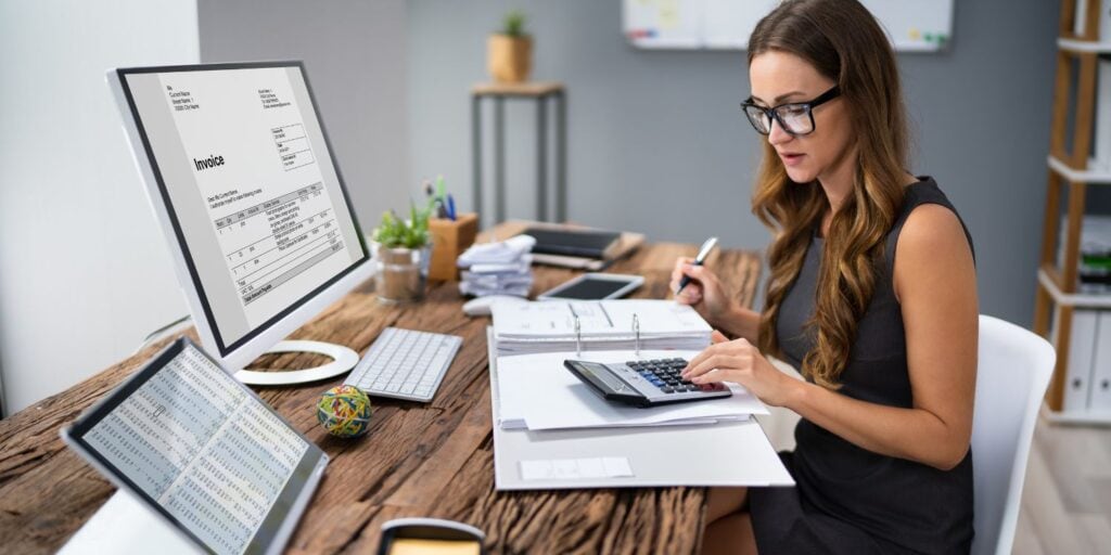 A small business owner doing her own accounting at her desk. She has a digital invoice on her computer screen and a big folder full of paper, and is using a calculator.