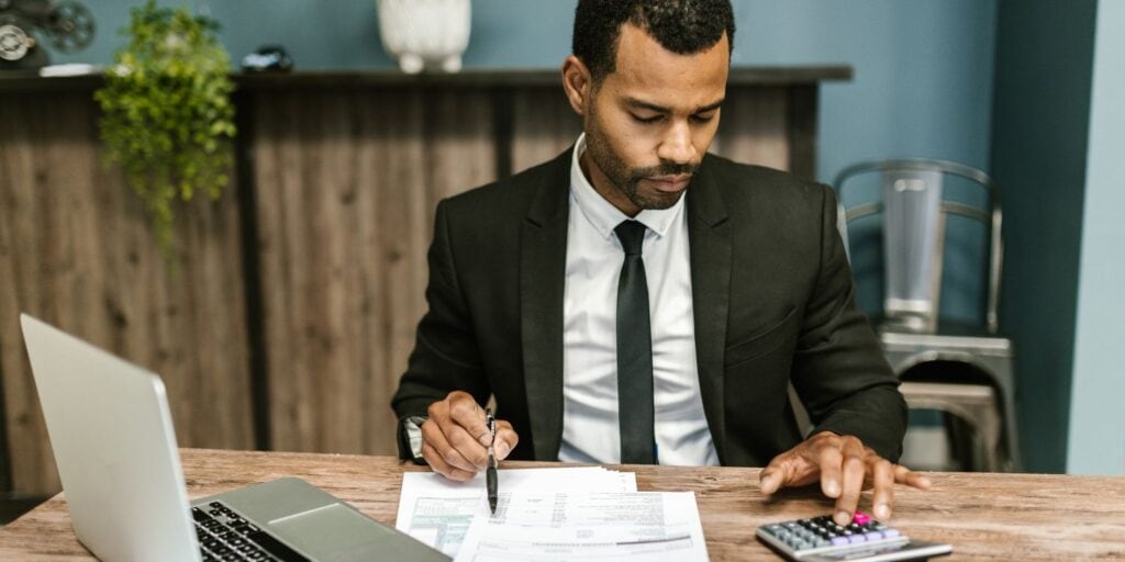 Man in a suit reviewing tax documents and using a calculator next to an open laptop, checking his tax return.